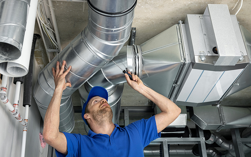 A repair man reaches over his head to inspect and update air ducts