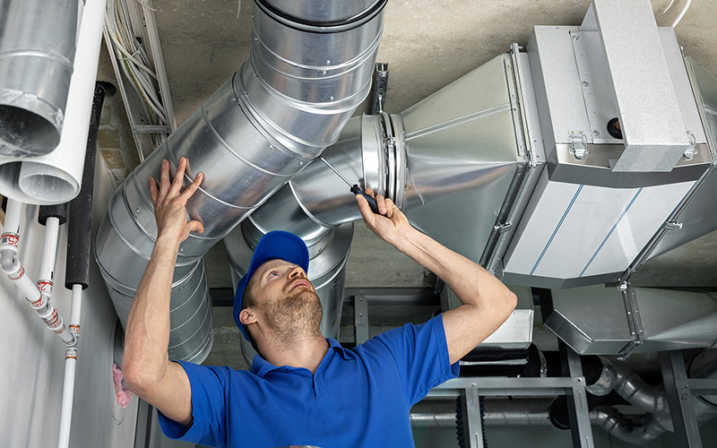 A repair man reaches over his head to inspect and update air ducts